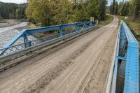 a metal bridge crosses over a dirt road near trees and a river in a remote region