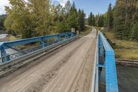 a metal bridge crosses over a dirt road near trees and a river in a remote region