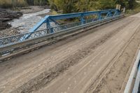 a metal bridge crosses over a dirt road near trees and a river in a remote region