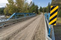 a metal bridge crosses over a dirt road near trees and a river in a remote region