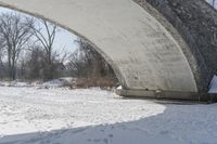 snow covers a road bridge in the wintertime with footprints in the snow, a bench and trees