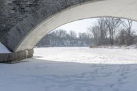 this stone bridge is over a snowy river near some trees and shrubs with footprints on the snow