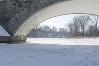 this stone bridge is over a snowy river near some trees and shrubs with footprints on the snow