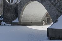 a snow covered stone arch under a bridge with snow around it and the ground with tracks on it