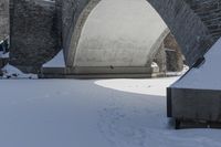 a snow covered stone arch under a bridge with snow around it and the ground with tracks on it