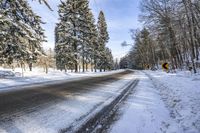 a snow covered road lined with trees next to some bushes and snowy ground is under blue sky