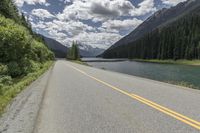 a road by the river leading up to mountains and some pine trees on both sides