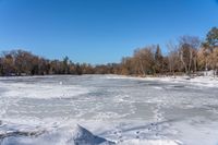 a body of water with trees in the background and snow on the ground and frozen water