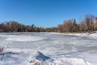 a body of water with trees in the background and snow on the ground and frozen water