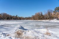 a body of water with trees in the background and snow on the ground and frozen water