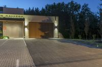 a lighted garage with plants on the side and a driveway leading into the entrance of an art gallery at night