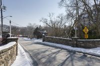 an empty street is lined with snow on the ground next to some buildings and a small stone fence