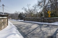 an empty street is lined with snow on the ground next to some buildings and a small stone fence