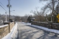 an empty street is lined with snow on the ground next to some buildings and a small stone fence