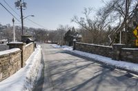 an empty street is lined with snow on the ground next to some buildings and a small stone fence