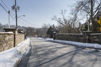 an empty street is lined with snow on the ground next to some buildings and a small stone fence