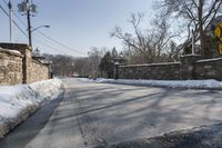 an empty street is lined with snow on the ground next to some buildings and a small stone fence