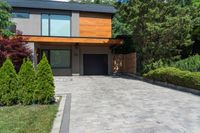 a brick paved driveway with shrubs and trees next to it, with a large wooden garage door and sliding glass doors