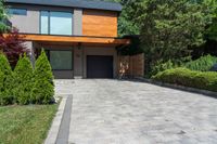 a brick paved driveway with shrubs and trees next to it, with a large wooden garage door and sliding glass doors