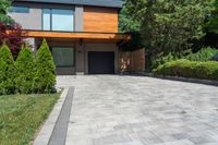 a brick paved driveway with shrubs and trees next to it, with a large wooden garage door and sliding glass doors