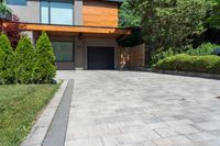 a brick paved driveway with shrubs and trees next to it, with a large wooden garage door and sliding glass doors