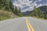 an empty mountain road and some trees and clouds in the sky and mountains and green grass