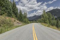 an empty mountain road and some trees and clouds in the sky and mountains and green grass