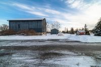 an older grey barn and a smaller dark one on a snowy road under a cloudy blue sky