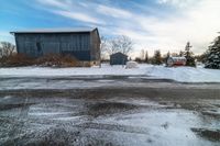 an older grey barn and a smaller dark one on a snowy road under a cloudy blue sky