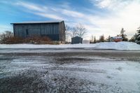 an older grey barn and a smaller dark one on a snowy road under a cloudy blue sky