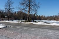 an empty city street covered in snow and ice next to a field of grass with trees and bushes