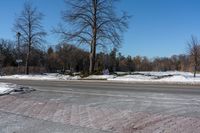 an empty city street covered in snow and ice next to a field of grass with trees and bushes