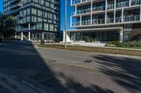 a street with an empty sidewalk and tall buildings in the background and one building has glass panels in the front and one has a glass bale windows