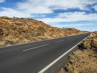 a paved road stretches along an arid terrain with mountains behind it and clouds in the sky