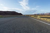 a paved road that is near a large hill with mountains in the background and a field on one side