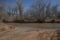 a dirt road in front of the river with trees and land behind it all in the foreground is a dirt patch and a creek