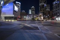 a street with buildings at night and a person riding a bike on the sidewalk by