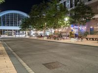 a road that leads to a shopping center at night with chairs and tables in the foreground