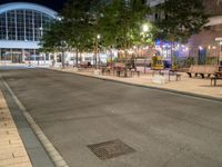 a road that leads to a shopping center at night with chairs and tables in the foreground