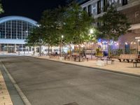 a road that leads to a shopping center at night with chairs and tables in the foreground