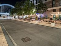 a road that leads to a shopping center at night with chairs and tables in the foreground