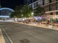 a road that leads to a shopping center at night with chairs and tables in the foreground