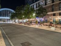 a road that leads to a shopping center at night with chairs and tables in the foreground