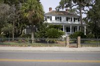 a house with white siding and a fence next to the street of a street lined by large trees