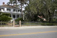 a house with white siding and a fence next to the street of a street lined by large trees