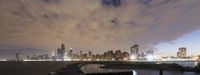 a bench sitting in front of some water near tall buildings with a cloudy sky above