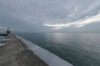 an overcast sky above the ocean as seen from a concrete dock with a lighthouse