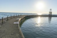 an empty pier next to the ocean with a sun shining above it and a lighthouse on a sunny day