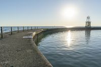 an empty pier next to the ocean with a sun shining above it and a lighthouse on a sunny day