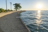 a concrete path leading over water near trees and grass near the ocean, with sun setting behind them